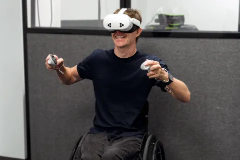 A male student in a wheelchair smiling while using a virtual-reality headset and two handheld controllers.