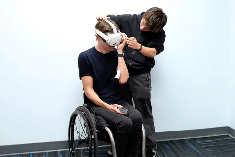 A male student  in a wheelchair wears a virtual-reality headset while another person adjusts it.