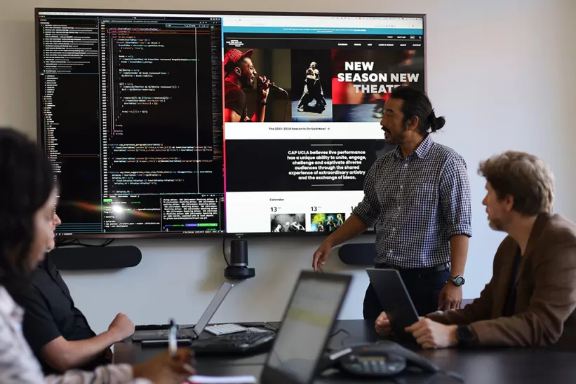 A group of four people sitting in a meeting room looking at a large display showing a coding interface and a theater webpage.