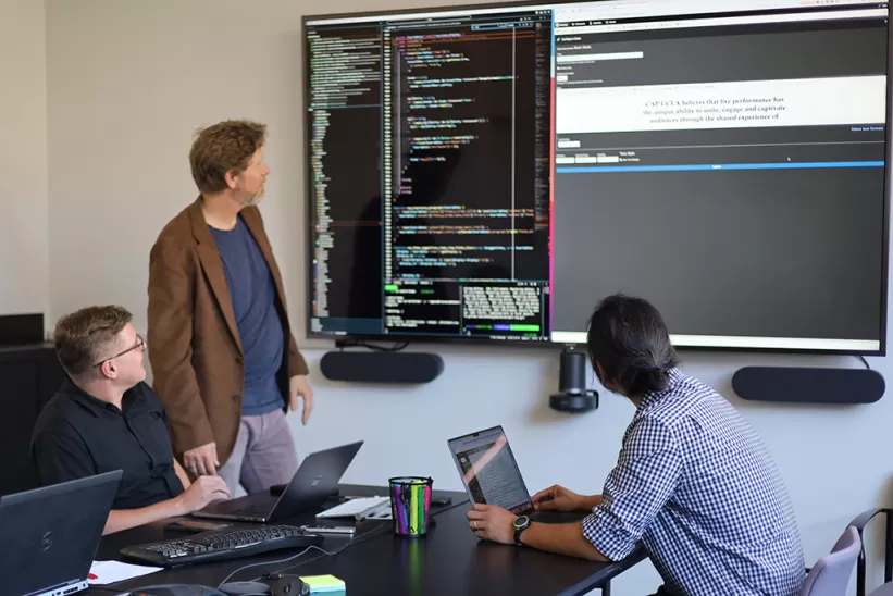 Three people in a meeting room with laptops out, sitting down at a conference table, looking at a large screen displaying code and a document.