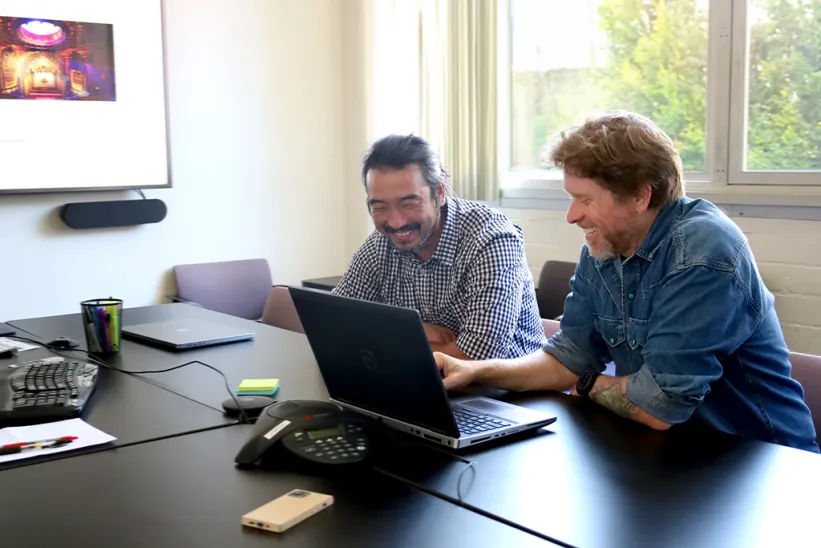 Two men talking at a table with a laptop in front of them.
