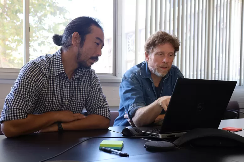 Two men seated at a table, looking at a laptop.