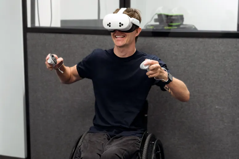 Smiling student in a wheelchair wearing a white VR headset and holds motion controllers, enjoying an immersive virtual reality experience indoors.
