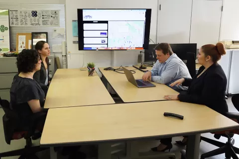 Four people seated at a table talking with a display screen behind them